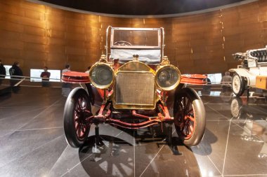 STUTTGART, GERMANY - OCTOBER 18, 2025: Front view of an early 20th-century antique brass era car with large round headlights and a radiator, displayed against a golden curved wall in the Mercedes-Benz Museum.
