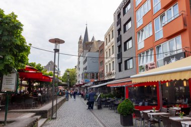 COLOGNE, GERMANY - 31 Mayıs 2024: Yayalar, Almanya 'nın tarihi Köln şehrinde tarihi bir kilise kulesi olan açık hava kafeleri ve restoranlarla dolu bir kaldırım taşı caddesi boyunca yürürler