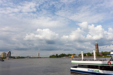 KOLOGNE, GERMANY - 31 Mayıs 2024: Cologne, Almanya 'da Ren nehrinin geniş bir manzarası, önplanda beyaz bir gezinti teknesi ve dramatik bulutlu bir gökyüzünün altında bir şehir silueti