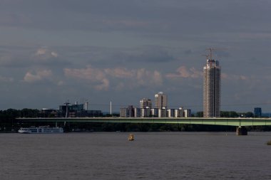 KOLOGNE, GERMANY - 31 Mayıs 2024: Cologne, Almanya 'daki Ren nehrinin panoramik manzarası, Zoobrucke köprüsü ve dramatik bulutlu gökyüzü altındaki uzun AXA Kulesi gökdeleni. Şehir nehir manzarası