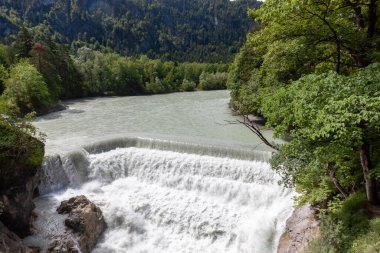 Fussen, Almanya 'da manzaralı Lechfall şelalesi. Güçlü turkuaz su, yazın yemyeşil orman ve Alp dağlarıyla çevrili çok katmanlı bir barajın üzerinden akıyor.