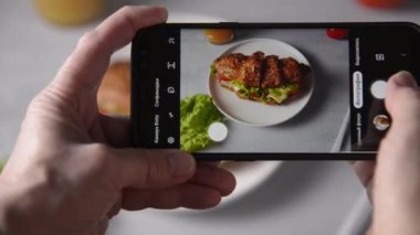 Kadın eli, kruvasan sandviçin akıllı telefonuyla et, sebze ve peynirli beyaz arka planlı fotoğraflarını çeker. Yemek fotoğrafı blogu kavramı.