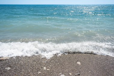 Kayalık çakıl taşı plajlı mavi deniz ve güneşli bir günde mavi gökyüzünün altında deniz köpüğüyle dalgalar ufuk çizgisi olan güzel doğa manzarası.