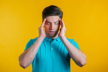 Baş ağrısı çeken genç bir adam, stüdyo portresi. Adam ellerini başının üstüne koyuyor, sarı arka planda izole edilmiş. Sorunlar, ilaç, hastalık kavramı