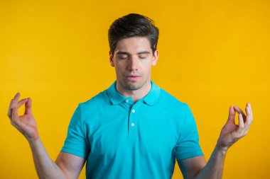 Mavi elbiseli sakin adam dinleniyor, meditasyon yapıyor. Sakinleşir, sarı stüdyo arka planında derin derin nefes alır. Yoga konsepti.