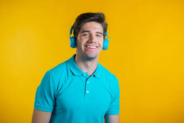 Jovem bonito com penteado na moda se divertindo, sorrindo, dançando com fones de ouvido azuis em estúdio contra fundo amarelo. Música, dança, conceito de rádio. — Fotografia de Stock