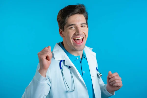 Médecin souriant danse drôle, succès et chance au travail. Jeune beau doc homme en manteau médical professionnel blanc est isolé sur fond de studio bleu. — Photo