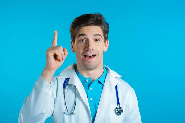 Porträt eines Arztes im Arztkittel, der auf blauem Studiohintergrund mit erhobenem Zeigefinger zeigt. Glücklich lächelnder Doktor zeigt Eureka-Geste. — Stockfoto