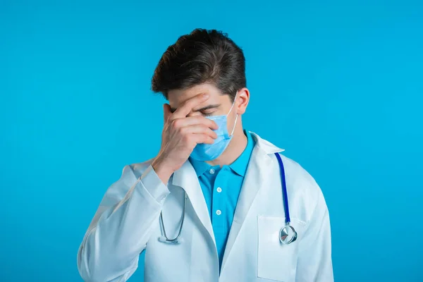 Sfrustrowany lekarz w białym ubraniu i masce na tle niebieskiej ściany. Doc jest zmęczony, znudzony pracą, rozczarowany, bezradny — Zdjęcie stockowe