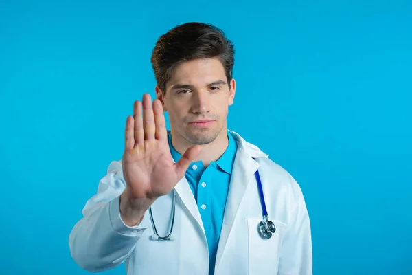 Portret poważnego lekarza w białym, medycznym płaszczu, wykazujący odrzucenie gestu znakiem stopu dłoni. Doc Man odizolowany na niebieskim tle. — Zdjęcie stockowe