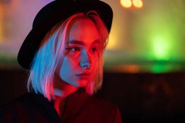 Neon parlak arka planda duran sarı saçlı hippi kadın. Geceleri şehir. Şapka, burun piercing 'i. Güzel, çekici kız..