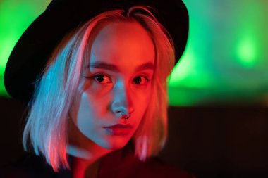 Neon parlak arka planda duran sarı saçlı hippi kadın. Geceleri şehir. Şapka, burun piercing 'i. Güzel, çekici kız..