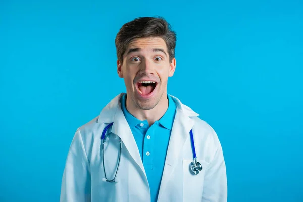 Şaşırmış doktor adam, vay canına diyor. Tıp önlüklü yakışıklı doktor mavi arka planda kameraya şaşırdı.. — Stok fotoğraf