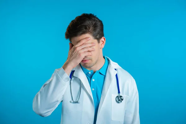 Απογοητευμένος γιατρός με λευκή φθορά πάνω από το μπλε φόντο στον τοίχο. Ο γιατρός είναι κουρασμένος, βαριέται τη δουλειά, απογοητεύεται, είναι αβοήθητος. — Φωτογραφία Αρχείου