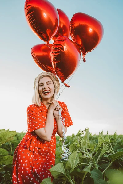 Ritratto di donna attraente in abito rosso in posa con palloncini a forma di cuore su sfondo verde natura. Ragazza con cappello vintage di paglia o berretto. Compleanno, vacanza, celebrare il concetto di libertà. — Foto Stock