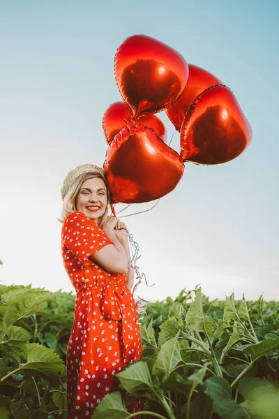 Portré vonzó nő piros ruhában pózol szív alakú lufik zöld természet háttér. Szalmakalapot vagy sapkát viselő lány. Születésnap, ünnep, szabadság ünnepe. — Stock Fotó