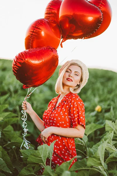 Portret atrakcyjnej kobiety w czerwonej sukience pozującej z balonami w kształcie serca na zielonym tle natury. Dziewczyna w słomkowym klasycznym kapeluszu lub berecie. Urodziny, wakacje, świętować koncepcję wolności. — Zdjęcie stockowe