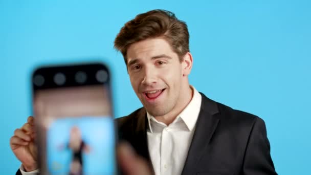 Sorrindo vlogger homem de negócios gravação de vídeo de si mesmo dançando na frente da câmera do smartphone no fundo azul. Gerente de terno faz clipe engraçado de mídia social — Vídeo de Stock