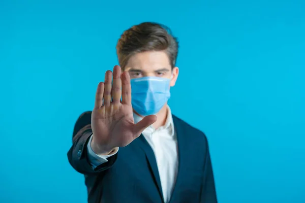 Retrato de hombre de negocios serio en traje profesional y máscara médica que muestra gesto de rechazo por señal de mano parada. Hombre aislado sobre fondo azul. — Foto de Stock