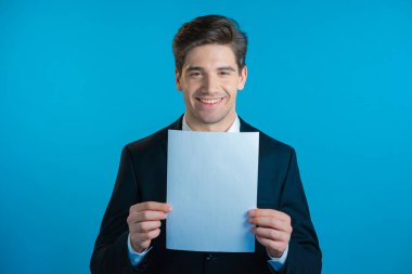 Takım elbiseli genç yakışıklı işadamının portresi elinde mavi stüdyo arka planında izole edilmiş beyaz bir a4 kağıt tutuyordu. Boşluğu kopyala.