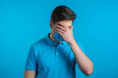 Maskeli genç bir adamın facepalm hareketi yaparken portresi, hayır, unuttum. Mutsuz adam üzüntü, pişmanlık, drama, başarısızlık hissediyor. Mavi duvarda izole edilmiş.