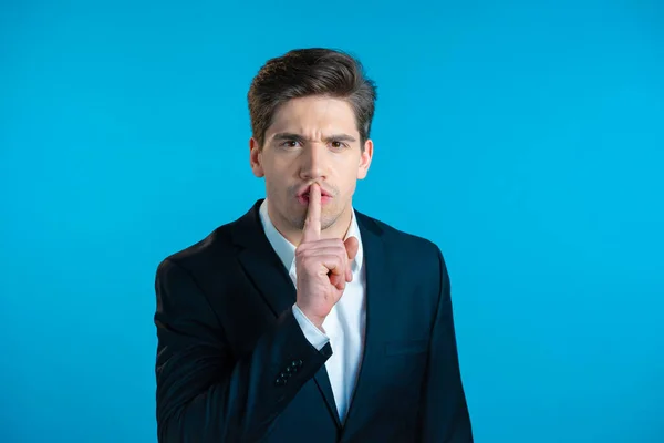 Serious business man in professional suit holding finger on lips over blue background. Gesture of shhh, secret, silence. Close up.