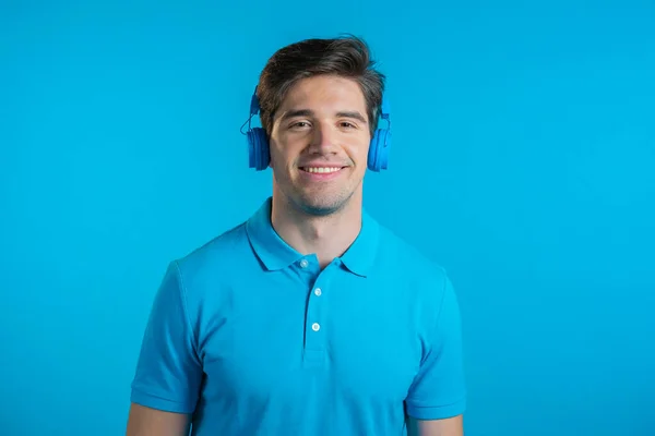 Beau jeune homme à la coiffure tendance s'amusant, souriant avec des écouteurs en studio sur fond bleu. Musique, danse, concept radio. — Photo