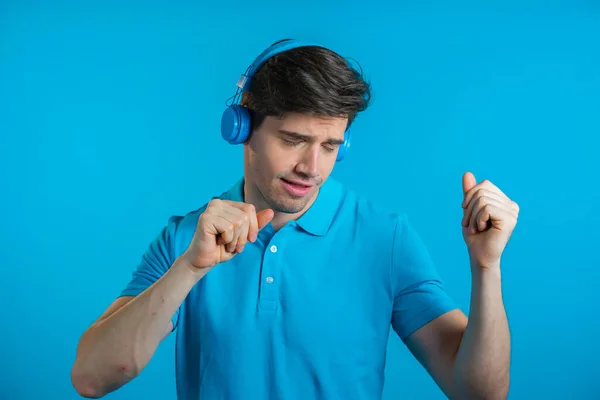 Jovem bonito com penteado na moda se divertindo, dançando, sorrindo com fones de ouvido em estúdio contra fundo azul. Música, dança, conceito de rádio. — Fotografia de Stock