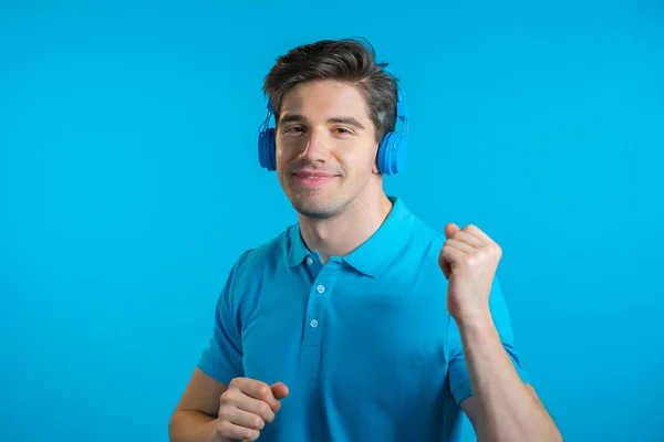 Beau jeune homme à la coiffure tendance s'amusant, souriant avec des écouteurs en studio sur fond bleu. Musique, danse, concept radio. — Photo