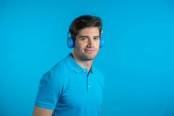 Beau jeune homme à la coiffure tendance s'amusant, souriant avec des écouteurs en studio sur fond bleu. Musique, danse, concept radio. — Photo