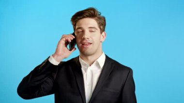 Genç işadamı telefonda gülümseyerek konuşur. Adam elinde tutuyor ve akıllı telefon kullanıyor. Takım elbise. Mavi stüdyo arka planı.