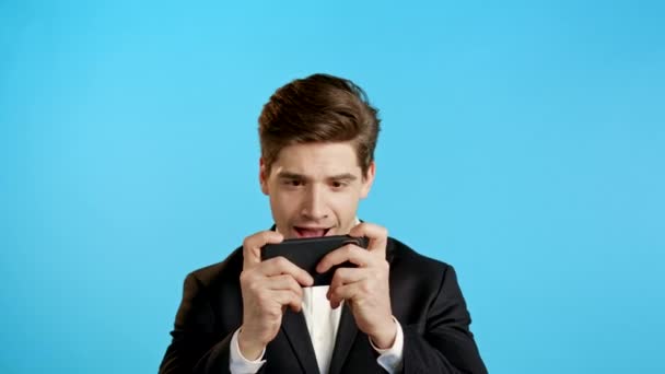 Beau homme d'affaires jouant au jeu sur smartphone sur mur studio bleu. Utilisation de la technologie moderne - applications, réseaux sociaux. — Video