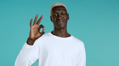 Melez yarışçı mavi arka planda baş parmak işareti gösteriyor. Pozitif Afrikalı adam kameraya gülümsüyor. Kazandım. Başarı. Vücut dili.