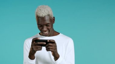 Mavi stüdyo duvarında akıllı telefondan oyun oynayan yakışıklı bir Afrikalı. Modern teknolojiyi kullanarak - uygulamalar, sosyal ağlar.