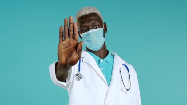 Portrait of serious black doctor in professional medical white coat and mask showing rejecting gesture by stop palm sign. Doc man isolated on blue background. — Stock Video