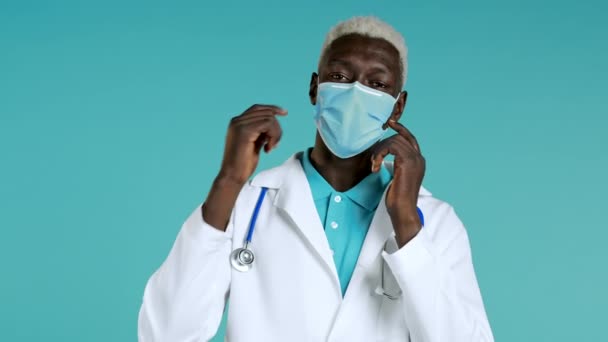 El doctor africano satisfecho con el abrigo médico quitó la máscara protectora como símbolo del fin pandémico y epidémico. Fondo estudio azul. — Vídeos de Stock