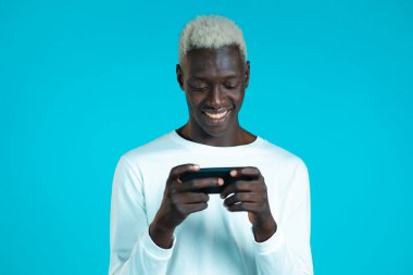 Mavi stüdyo duvarında akıllı telefondan oyun oynayan yakışıklı bir Afrikalı. Modern teknolojiyi kullanarak - uygulamalar, sosyal ağlar.