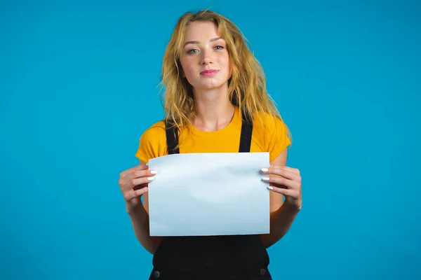 Retrato de una joven mujer seria sosteniendo papel a4 blanco aislado sobre fondo de estudio azul.Espacio de copia. —  Fotos de Stock