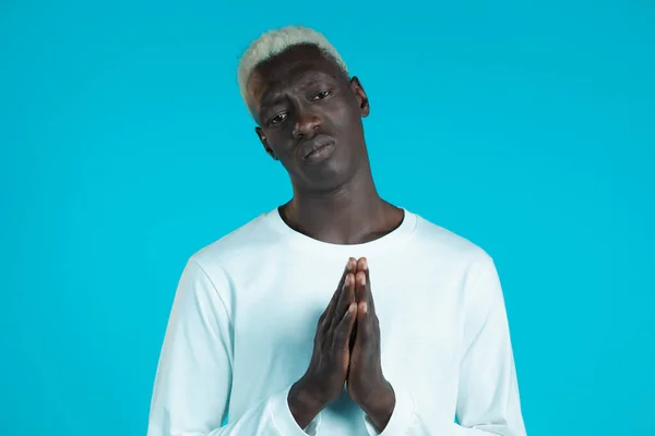 Joven africano de manga larga blanca rezando sobre fondo azul. Chico rogando a alguien satisfacer sus deseos, ayuda con. — Foto de Stock