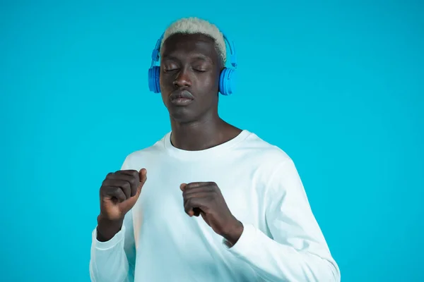 Hombre negro guapo con peinado de moda divirtiéndose, sonriendo, bailando con auriculares en el estudio sobre fondo azul. Música, danza, concepto de radio. — Foto de Stock