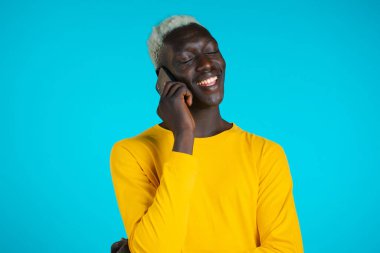 Genç Afrikalı Amerikalı adam telefonda gülümseyerek konuşuyor. Adam elinde tutuyor ve akıllı telefon kullanıyor. Mavi stüdyo arka planı.