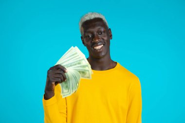 Parası olan mutlu Afrikalı bir adam mavi stüdyo duvarında Amerikan doları banknotları. Başarı, kazanç, zafer sembolü.