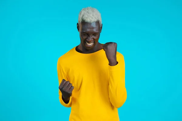 Homem afro-americano em amarelo é muito feliz e feliz, ele mostra sim gesto de vitória, cara alcançado resultado, objetivos. Surpreendido animado menino feliz no fundo azul — Fotografia de Stock