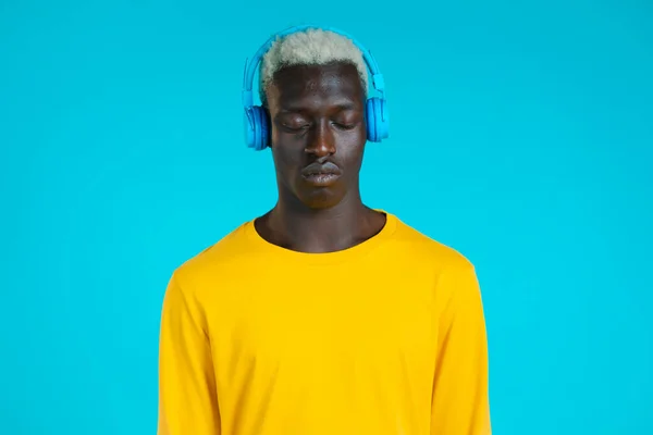 Hombre negro guapo con peinado de moda y auriculares en el estudio sobre fondo azul. Un tipo de traje amarillo. Música, danza, concepto de radio. — Foto de Stock