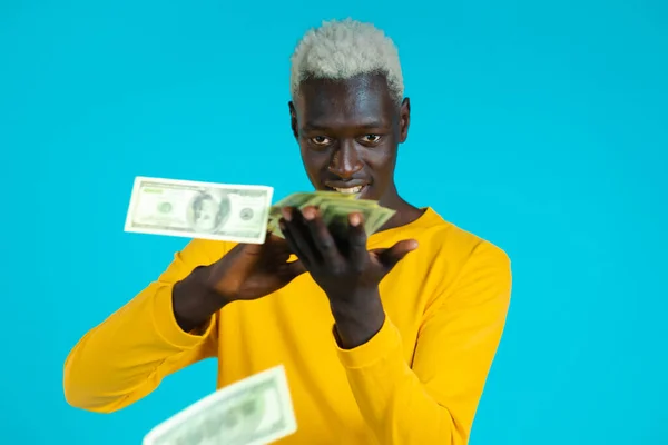 Hombre de raza mixta con cara feliz dispersa el dinero. La gente gasta en exceso la moneda estadounidense. Africano hombre es rubor con dólares en azul estudio fondo. — Foto de Stock