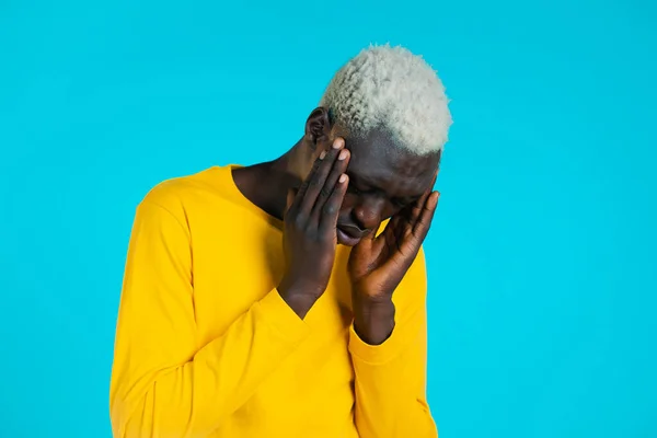 Joven africano de amarillo con dolor de cabeza, retrato de estudio. Un tipo poniendo las manos en la cabeza, aislado sobre fondo azul. Concepto de problemas, medicina, enfermedad — Foto de Stock