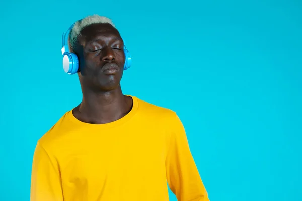 Bel homme noir avec coiffure tendance et écouteurs en studio sur fond bleu. Un type en tenue jaune. Musique, danse, concept radio. — Photo