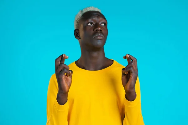 Joven africano de manga larga y amarilla rezando sobre fondo azul. Chico rogando a alguien satisfacer sus deseos, ayuda con. — Foto de Stock