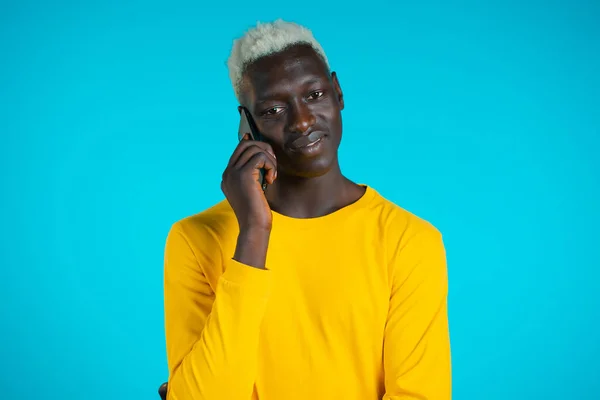 Jovem afro-americano fala com sorriso no telefone. Um tipo a segurar e a usar um telemóvel inteligente. Azul estúdio fundo. — Fotografia de Stock