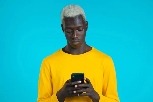 青いスタジオの壁にスマートフォンを使っているアフリカ人のハンサムな男。現代の技術を使用する-アプリ、ソーシャルネットワーク. — ストック写真